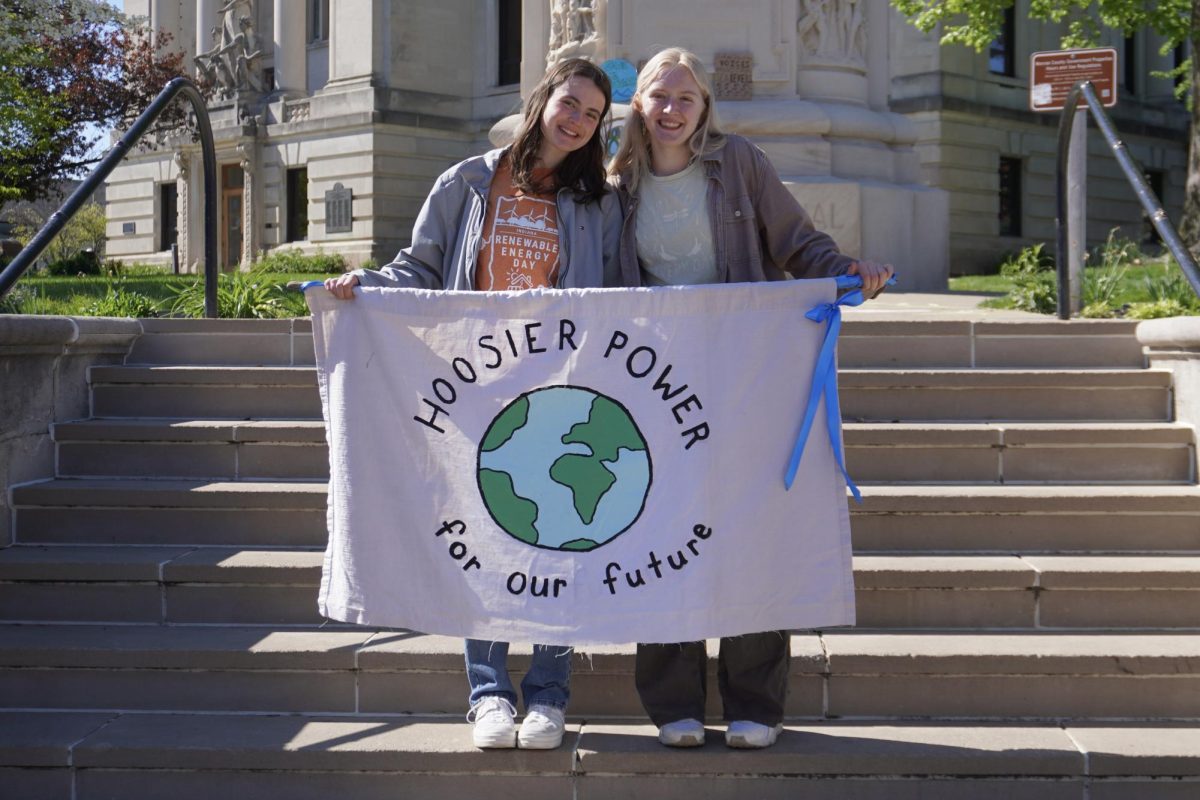 Organizers Alice Racek (left) and Lydia Arnold (right) hold a banner at the Hoosier Power Energy Rally in Bloomington